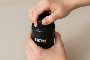 サーモスの黒い水筒のふたを開けようと指で押している様子。白木のテーブルに置かれたボトルを両手でしっかり支えている。