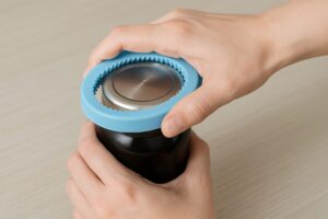 青いゴム製の開栓補助器具を使って、瓶の金属キャップを回して開けようとしている様子。テーブルの上に置かれた瓶と両手が写っている。
