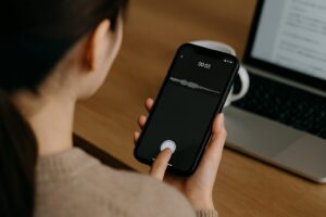 白いイヤホンを着けた女性がスマートフォンの音声アプリを操作している様子。背景にはノートパソコンと観葉植物が写っている。