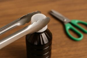 キッチン用トングで白いボトルキャップをつかんでいる様子。背景には木のテーブルと緑のはさみが置かれている。